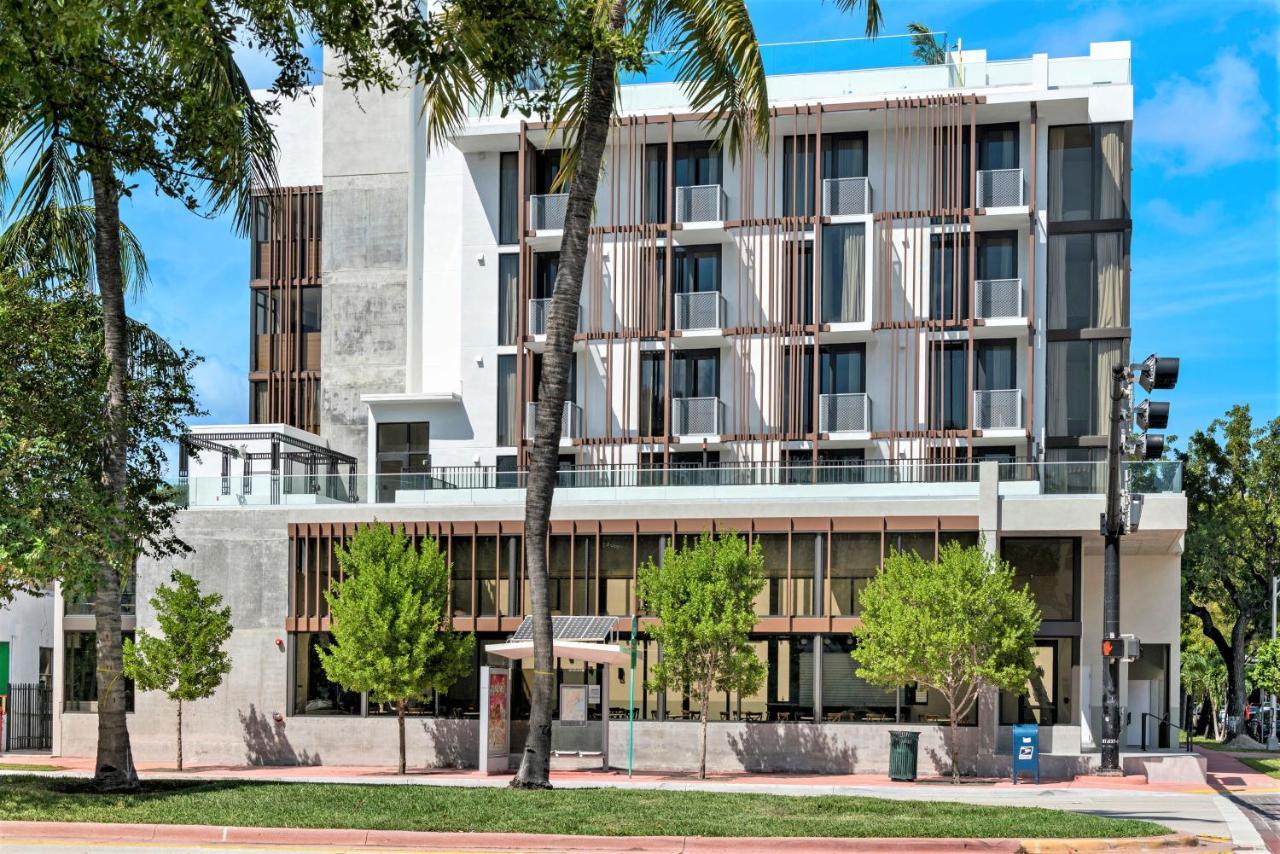Street-level view of Upsun Hotel facade with brown aluminum louvers on upper floors and ground-floor storefront system with palm trees - Miami Beach FL