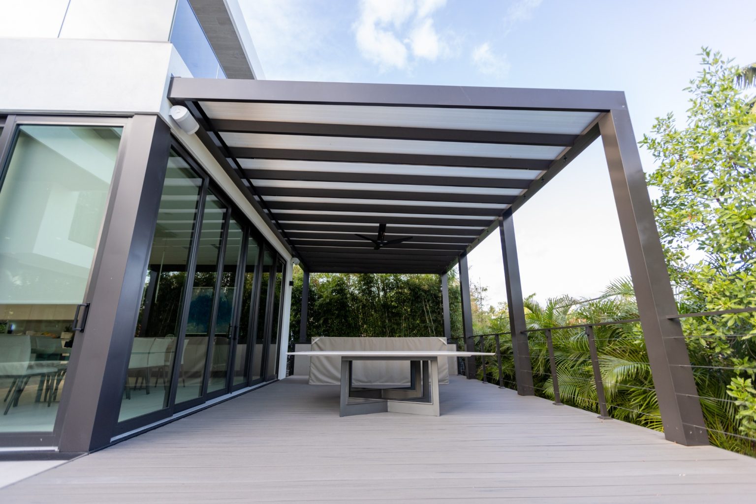 Covered pool deck with modern pergola and cable railing overlooking lush greenery at North Bay Village residence in Miami